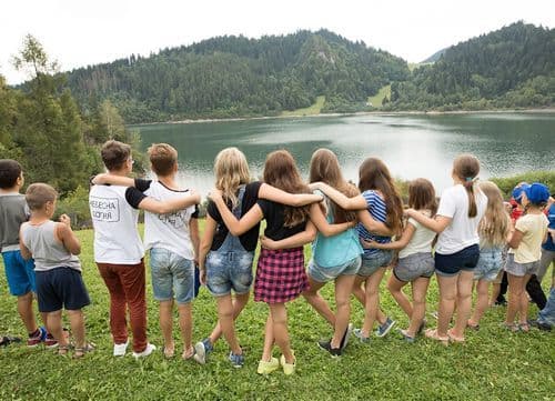 Children with their arms on each others shoulders looking at a lake 